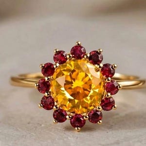 Puede incluir: Un anillo de oro con una gran gema amarilla central rodeada de pequeñas gemas rojas. El anillo tiene una fina banda dorada. La imagen está sobre un fondo neutro. El texto "London Jewellers" es visible en la parte superior.