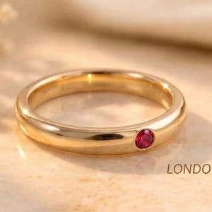 Könnte beinhalten: Ein polierter Goldring mit einem einzelnen, kleinen, runden und leuchtend roten Rubin, der bündig in das Metall eingelassen ist. Der Text LONDONJEWELLERS ist oben und unten rechts im Bild zu sehen und rahmt das Schmuckstück ein.