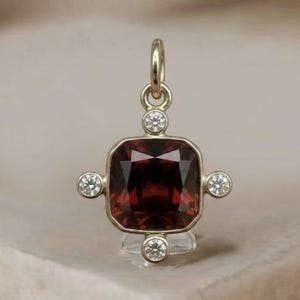 May include: A gold pendant with a square, deep red gemstone at its center, surrounded by four smaller, round, clear gemstones. The pendant has a loop at the top for a chain. The text "London Jewellers London Jewellers" is at the top.