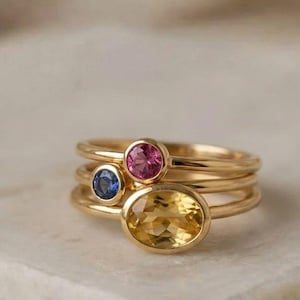 May include: Three gold rings with gemstones. The top ring has a pink stone, the middle ring a blue stone, and the bottom ring a yellow oval stone. The rings are stacked on a white surface. The text "London Jewellers" is at the top.