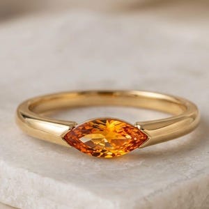 May include: A gold ring featuring an orange marquise-cut gemstone. The ring is displayed on a white surface. The words "London Jewellers" are at the top of the image.