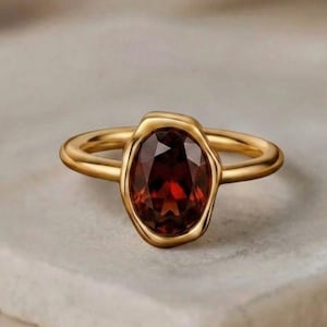 May include: A gold ring featuring a central oval-cut deep red garnet gemstone set within an irregular, organic-shaped gold bezel. The text London Jewellers is visible at the top of the frame. The band is smooth and circular in design.