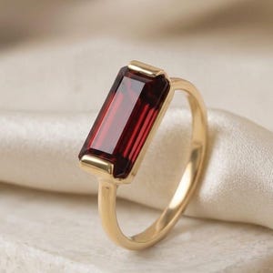 May include: A gold ring featuring a rectangular, deep red gemstone. The ring has a simple, elegant design with a thin band and a bezel setting. The gemstone is the focal point, showcasing its rich color and clarity. The ring is displayed on a white surface.