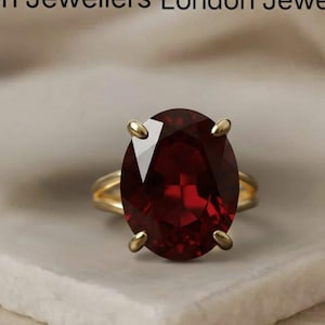 May include: A gold ring with a large, oval-shaped, deep red gemstone. The ring has a classic design with four prongs holding the stone. The words "London Jewellers" are at the top of the image.