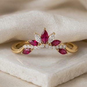 May include: A gold ring with a crown design, featuring marquise-cut ruby and diamond gemstones. The ring has a curved band and is set on a white surface. The words "London Jewellers" are at the top of the image.