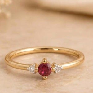 May include: A gold ring featuring a central round-cut red gemstone flanked by two small clear stones on each side. The band is thin and polished. The text LONDONJEWELLERS is visible at the top of the image. The design is simple and symmetrical.