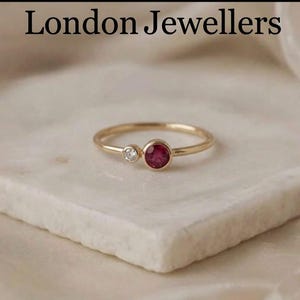 May include: A gold ring with a small, round, red gemstone and a smaller, clear gemstone. The ring is set on a white surface. The text "London Jewellers" is at the top of the image.
