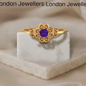 May include: A gold-colored ring with a floral design and a round, purple gemstone at the center. The ring is displayed on a white, textured surface. The words "London Jewellers" are at the top of the image.