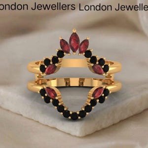 Puede incluir: Un anillo dorado con un diseño de doble banda. El anillo está adornado con piedras preciosas rojas y negras. La parte superior del anillo presenta tres piedras rojas en forma de marquesa, mientras que los lados y la parte inferior tienen una serie de piedras negras.