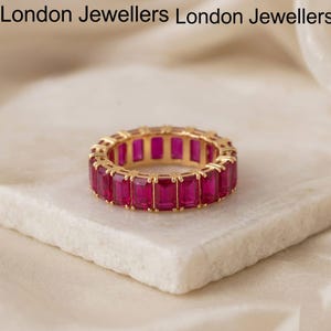 Könnte beinhalten: Ein goldfarbener Eternity-Ring mit rechteckigen, leuchtend rosa Edelsteinen. Der Ring liegt auf einer weißen, strukturierten Oberfläche. Die Worte "London Jewellers" sind oben im Bild aufgedruckt.