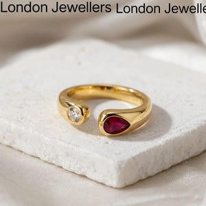 May include: A gold open-style ring featuring a round clear stone on one end and a pear-shaped red stone on the other. The text London Jewellers is visible at the top of the frame. The ring has a smooth, polished finish and a bypass design.