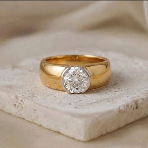 Puede incluir: Un anillo de oro con una piedra preciosa blanca de talla brillante redonda engastada en un bisel liso. La banda es ancha y pulida, mostrando un acabado metálico de alto brillo que contrasta con la piedra clara facetada en el centro.