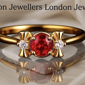Op de afbeelding: Een gouden ring met een centrale ronde rode edelsteen geflankeerd door twee kleinere ronde heldere stenen, met de tekst London Jewellers London Jewellers erboven. De ringband is van gepolijst goud en de zetting heeft decoratieve metalen accenten.