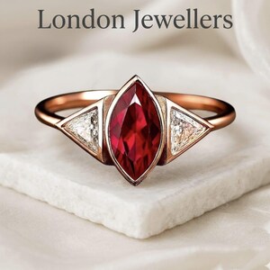 May include: A rose gold ring featuring a central marquise-cut deep red gemstone flanked by two triangular clear stones. The text London Jewellers is displayed above the ring, which is set on a textured white surface with soft fabric folds.