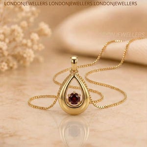May include: A gold pendant necklace featuring a teardrop-shaped frame with a single round dark red gemstone suspended in the center. The pendant hangs from a delicate gold box chain. The text LONDONJEWELLERS is visible at the top of the image.