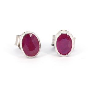 May include: A pair of silver stud earrings, each with an oval-cut ruby gemstone. The rubies are a deep red hue, set in a simple silver bezel. The earrings are presented against a white backdrop, highlighting the gemstones' colour and the silver setting.