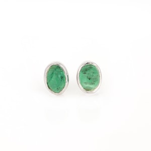 May include: A pair of oval-shaped stud earrings. Each earring features a vibrant green gemstone set in a silver-colored metal frame. The gemstones have a natural, textured appearance, and the metal frames have a smooth, polished finish.