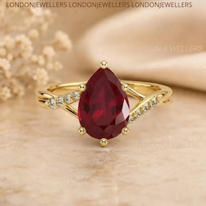 May include: A gold ring featuring a large pear-cut deep red gemstone at the centre, flanked by two split bands encrusted with small clear stones. The text LONDONJEWELLERS is visible in the background. The design showcases a polished metal finish.