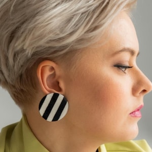 以下が含まれることがあります： 短いブロンドの髪をした女性が、ライムグリーンのジャケットを着ています。ジャケットには黒と白のストライプの円形のピンが付いています。彼女はまた、黒と白のストライプの円形の大きなイヤリングをしています。