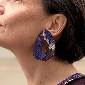 Può includere: Una donna indossa un maglione viola, una collana abbinata e grandi orecchini a goccia. I gioielli presentano un motivo a pelle di serpente blu, viola e bianco. Gli orecchini hanno un design ritagliato.