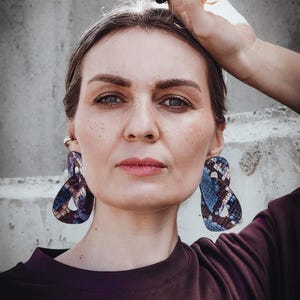 Könnte beinhalten: Eine Person trägt große, abstrakt geformte Ohrringe mit einem blau-braun-weißen Muster. Die Ohrringe sind der Blickfang, ergänzt durch ein dunkelviolettes Oberteil und einen Ring an der Hand.