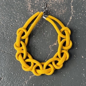 Puede incluir: Un collar de cuerda amarillo con un diseño anudado. El collar está hecho de cuerda gruesa trenzada y tiene un cierre plateado.