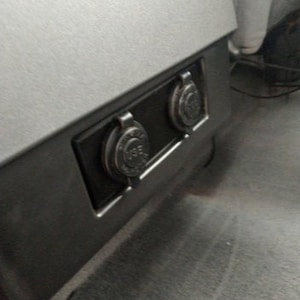 Może przedstawiać: Dwa czarne gniazdka elektryczne są zamontowane w szarym panelu plastikowym. Gniazdka są osadzone w panelu i mają czarną plastikową osłonę.