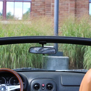 Op de afbeelding: Interieur van een zwarte cabriolet met bruin lederen stoelen en een houten stuurwiel. De achteruitkijkspiegel is zichtbaar en de auto staat geparkeerd voor een gebouw met ramen en hoog gras.