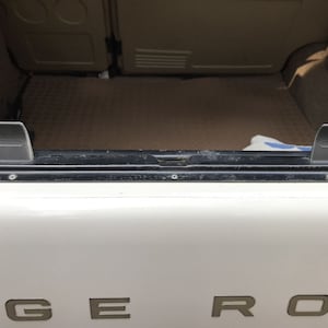 Può includere: Un'auto bianca con la scritta "RANGE ROVER" sul retro. Il portellone posteriore è aperto e ci sono due clip di plastica nera sul bordo del portellone.