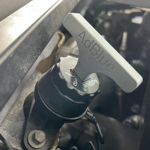 Puede incluir: Primer plano de un componente del motor de un coche. Una tapa gris AdBlue con un asa a juego se inserta en un componente negro. El texto "AdBlue" es visible en el asa y en la tapa. El fondo es un compartimento oscuro del motor.
