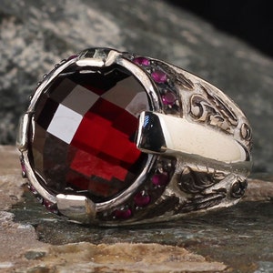 May include: A silver ring featuring a large, red gemstone surrounded by smaller, pink gemstones. The ring has an intricate design with a cross-shaped detail.