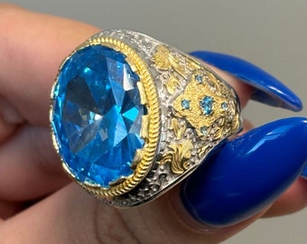 May include: A silver and gold ring with a large blue gemstone. The ring has intricate details and smaller blue gemstones around the main stone.