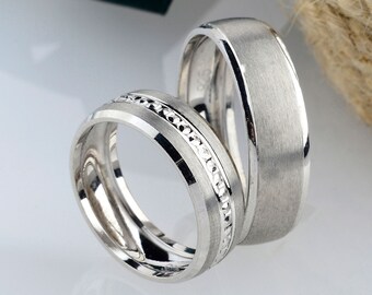May include: Two silver wedding bands, one with a row of small diamonds, on a white surface.