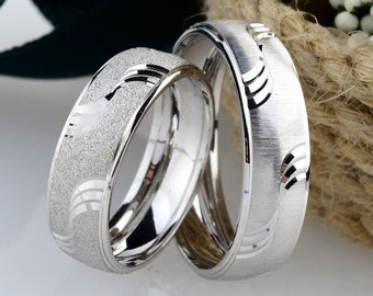 May include: Two silver wedding bands with a textured finish. The bands have a wave-like design and are displayed on a rustic background.