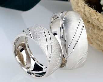May include: Two silver wedding bands with a textured and a grooved design. The bands are on a white surface with a green ribbon and a basket of white flowers in the background.