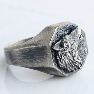 May include: A silver-toned ring featuring a detailed howling wolf design. The wolf's head is set within a textured, octagonal frame. The ring has a brushed metal finish, giving it a vintage or antique appearance. The ring is a piece of jewelry.