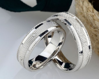 May include: Two silver wedding rings with textured bands and small diamond accents. The rings are intertwined, reflecting light. A dark green ribbon and a woven basket with white flowers are in the background.