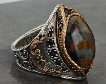May include: A silver ring with a large, oval-shaped tiger's eye gemstone set in a gold-toned bezel. The ring is decorated with intricate floral designs and black gemstones.