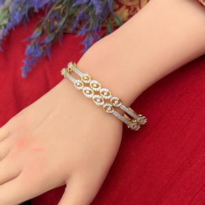 May include: A gold and silver-toned bracelet with a repeating oval design, adorned with small, clear gemstones. The bracelet is worn on a hand with manicured nails, set against a red fabric background. Purple flowers are visible in the background.
