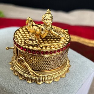 May include: A gold-colored, ornate box with a detailed lattice pattern and a red gemstone border. A small, gold figurine of a person is lying on top. The box has a decorative base and a small handle.