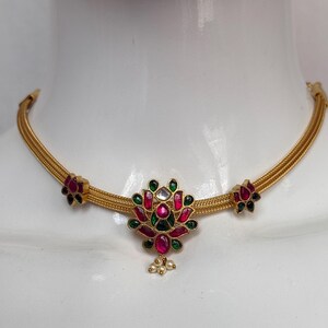 May include: A gold-toned necklace with a floral pendant featuring red, green, and white gemstones. The necklace has a delicate chain design and is displayed on a white mannequin. Additional floral accents are on each side.