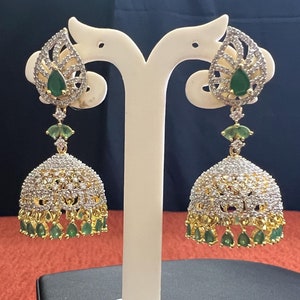 May include: Elegant gold-toned earrings with a teardrop design, adorned with clear and emerald-green gemstones. The earrings feature a bell-shaped bottom with dangling green gems and are displayed on a white stand, showcasing their intricate details.
