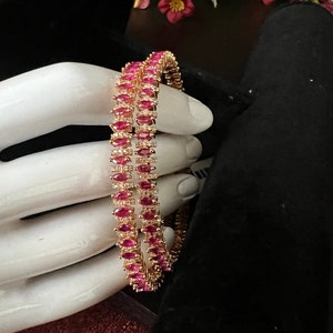 May include: Two gold bangles with pink marquise-shaped stones set in a row around the bangle. The bangles are on a white hand model.