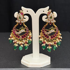 May include: Gold-toned chandelier earrings with ruby, emerald, and pearl accents. The earrings feature a crescent design with a central row of white stones and dangling green and red beads. The earrings are displayed on a white stand against a black background.