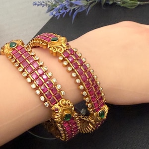 May include: Gold-toned bangles with a repeating pattern of square, ruby-colored stones and small clear stones. The bangles have ornate gold detailing with green and clear stones. The bangles are on a person's wrist.