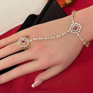 May include: A gold-toned hand chain with a ring and bracelet design. The jewelry features a floral design with a central red gemstone surrounded by clear crystals. The ring is on the ring finger, and the bracelet is on the wrist.