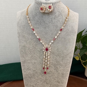 May include: A gold-toned necklace and earring set featuring clear pear-shaped stones and small oval-shaped red stones. The necklace has a central floral-inspired pendant with three dangling strands of stones, while the earrings are matching studs.