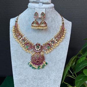May include: A gold-toned necklace and matching jhumka earring set featuring embedded red and green stones, clear crystals, and small dangling white pearls. The central pendant of the necklace displays a detailed floral motif with green accents.