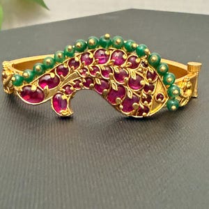 May include: Gold-toned bangle bracelet with a central design featuring ruby-colored stones and green beads. The bracelet has an ornate, floral-inspired pattern and a hinged clasp. The design is set against a dark background.