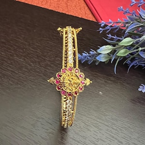 May include: A gold bangle bracelet with a floral design and pink and white stones. The bracelet is on a dark brown surface.